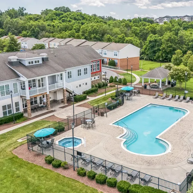 Apartment Complex Pool Area An aerial view of an apartment complex featuring a swimming pool and surrounding amenities. The pool is shaped irregularly and is enclosed by a black fence. There are lounge chairs lined by the pool, and a jacuzzi nearby. The complex includes multiple two-story buildings set against a backdrop of green trees and a well-maintained lawn.