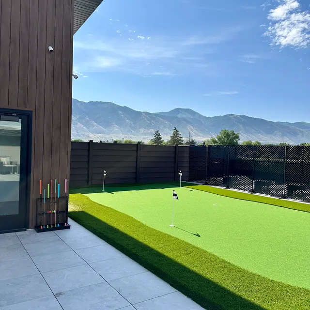 A backyard golf putting green surrounded by mountains and a wooden fence, with a clear blue sky and a modern building in the foreground.