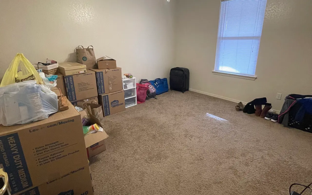 A spacious room with light beige carpet, a ceiling fan, and a window letting in natural light. Boxed items are stacked on one side, while bags and various items are scattered on the floor.