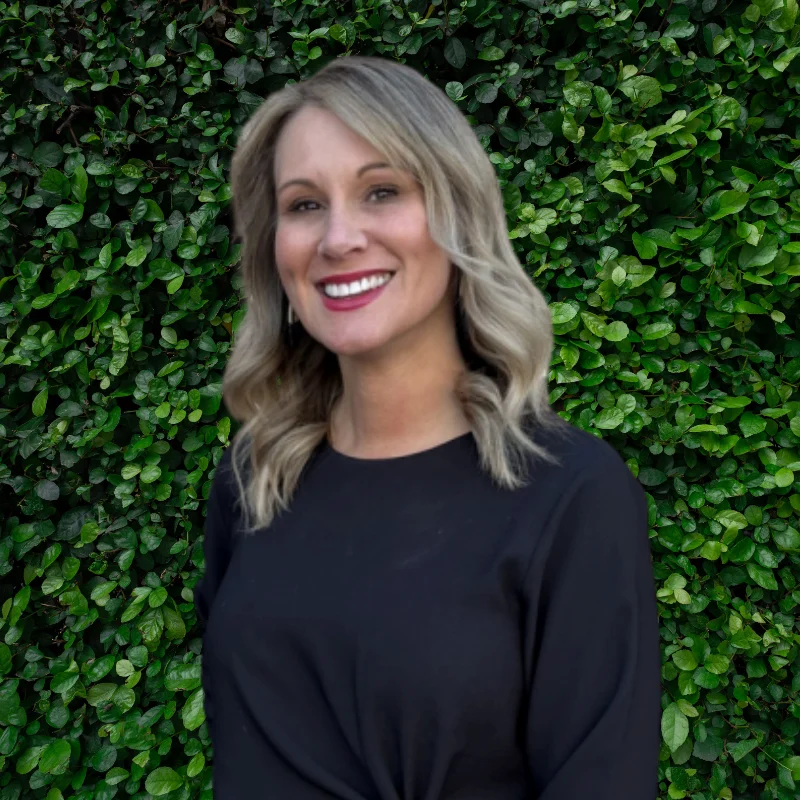 A smiling woman with medium-length blonde hair stands in front of a lush green wall. She is wearing a black top and is looking directly at the camera with a friendly expression.