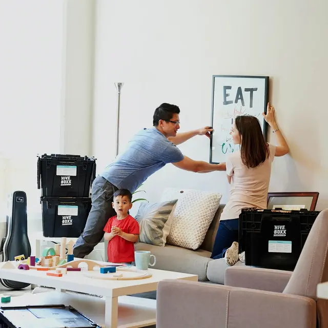 A family is in a living room setting with boxes scattered around. A man is hanging a sign on the wall while a woman assists him. A young child watches nearby. The room has a sofa, coffee table with toys, and several packing boxes labeled for moving.