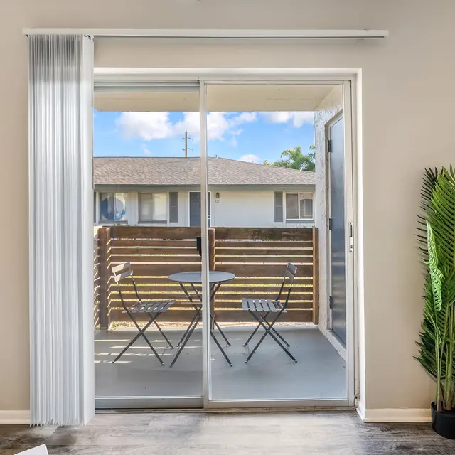 A cozy living space with glass sliding doors leading to a small balcony. The balcony features a table and two chairs, surrounded by wooden fencing. There's a green plant in the corner of the living space, and the interior has a neutral color scheme.