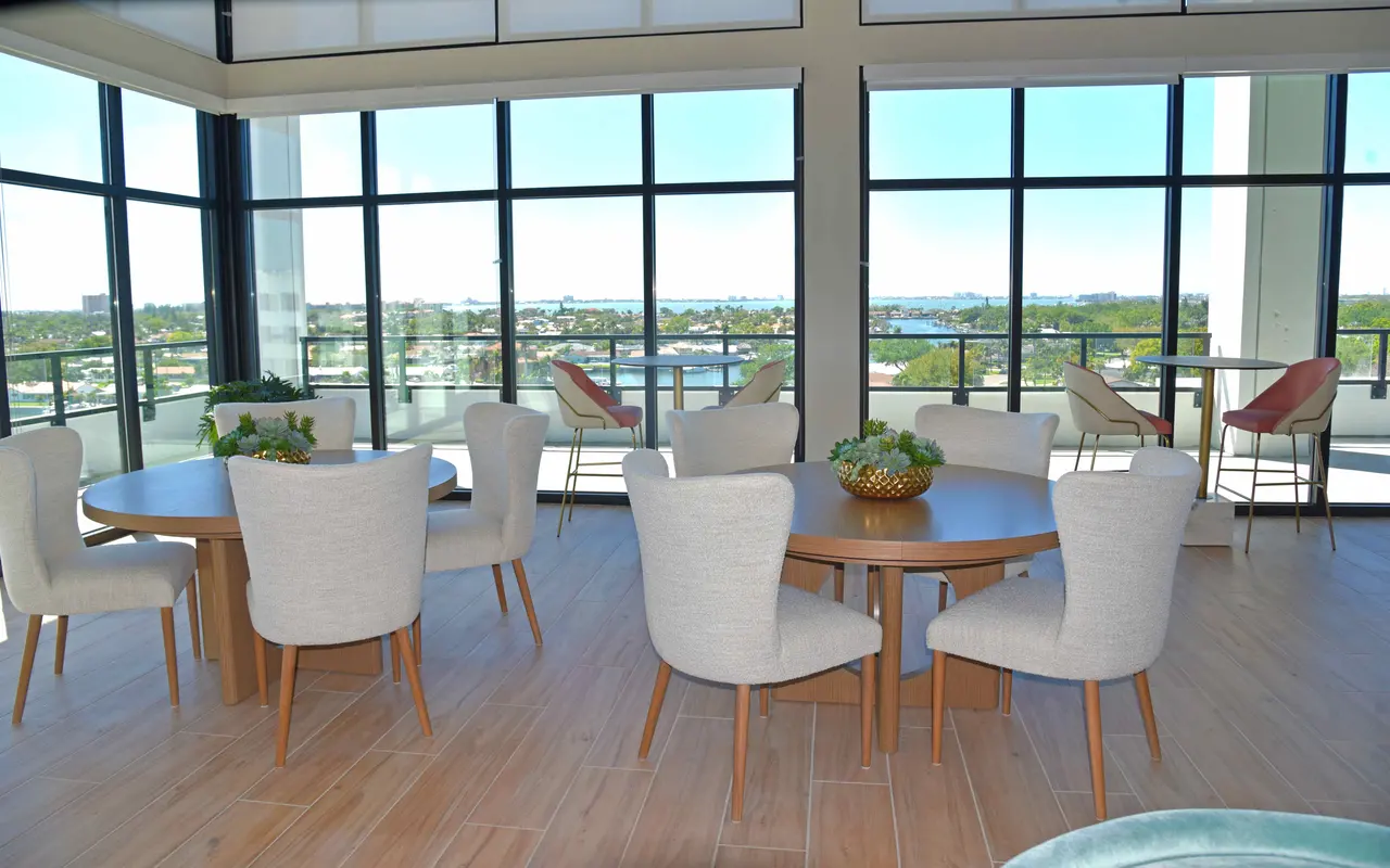 A modern meeting space with large windows offering a view of the outdoors. There are multiple round tables with chairs arranged in the center, and high chairs at a bar-like area near the windows.