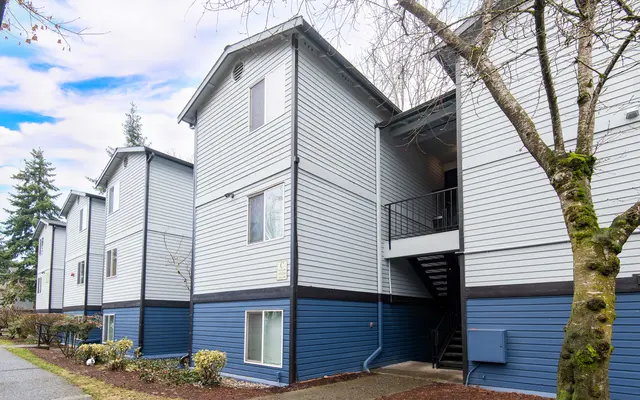 A multi-unit apartment building with blue and gray siding, featuring three floors, windows, and a staircase leading to an upper level. Trees are visible in the foreground.