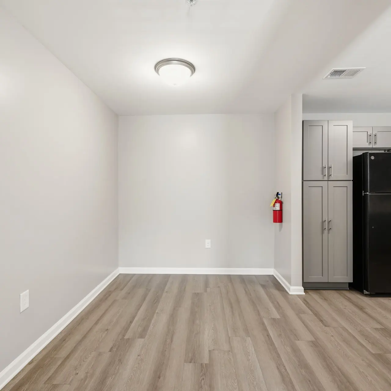 A spacious, unfurnished room with light gray walls and wood-like flooring. A black refrigerator is visible in the corner, next to gray cabinets, and a fire extinguisher mounted on the wall.