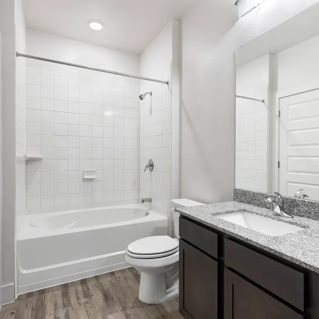 A modern bathroom featuring a bathtub with a shower, a toilet, and a double vanity with a granite countertop. The walls are tiled with white squares and the floor is covered with wooden planks. There is a large mirror above the vanity and bright overhead lighting.