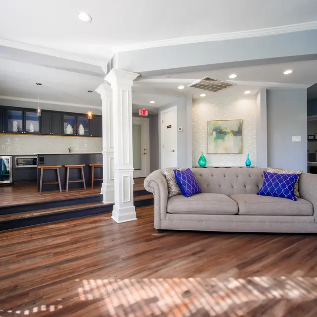 A spacious modern living room featuring a light gray tufted sofa with blue accents, wooden flooring, and a stylish open kitchen in the background.