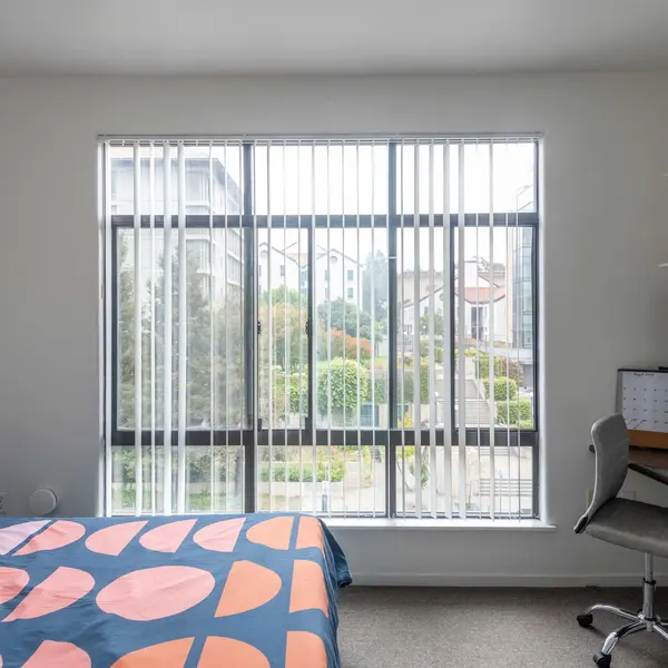 A modern bedroom with a bed covered in a pink and blue patterned blanket, a large window with vertical blinds showing a garden view, and a workspace with a desk and office chair.