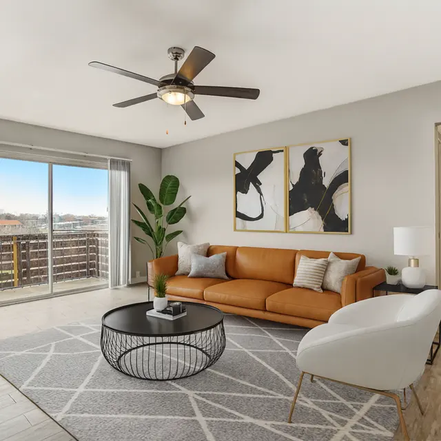 A modern living room featuring a brown leather sofa, white accent chair, and a circular coffee table on a geometrically patterned rug. There's a large window showing a scenic view, potted plants, and contemporary art on the walls.