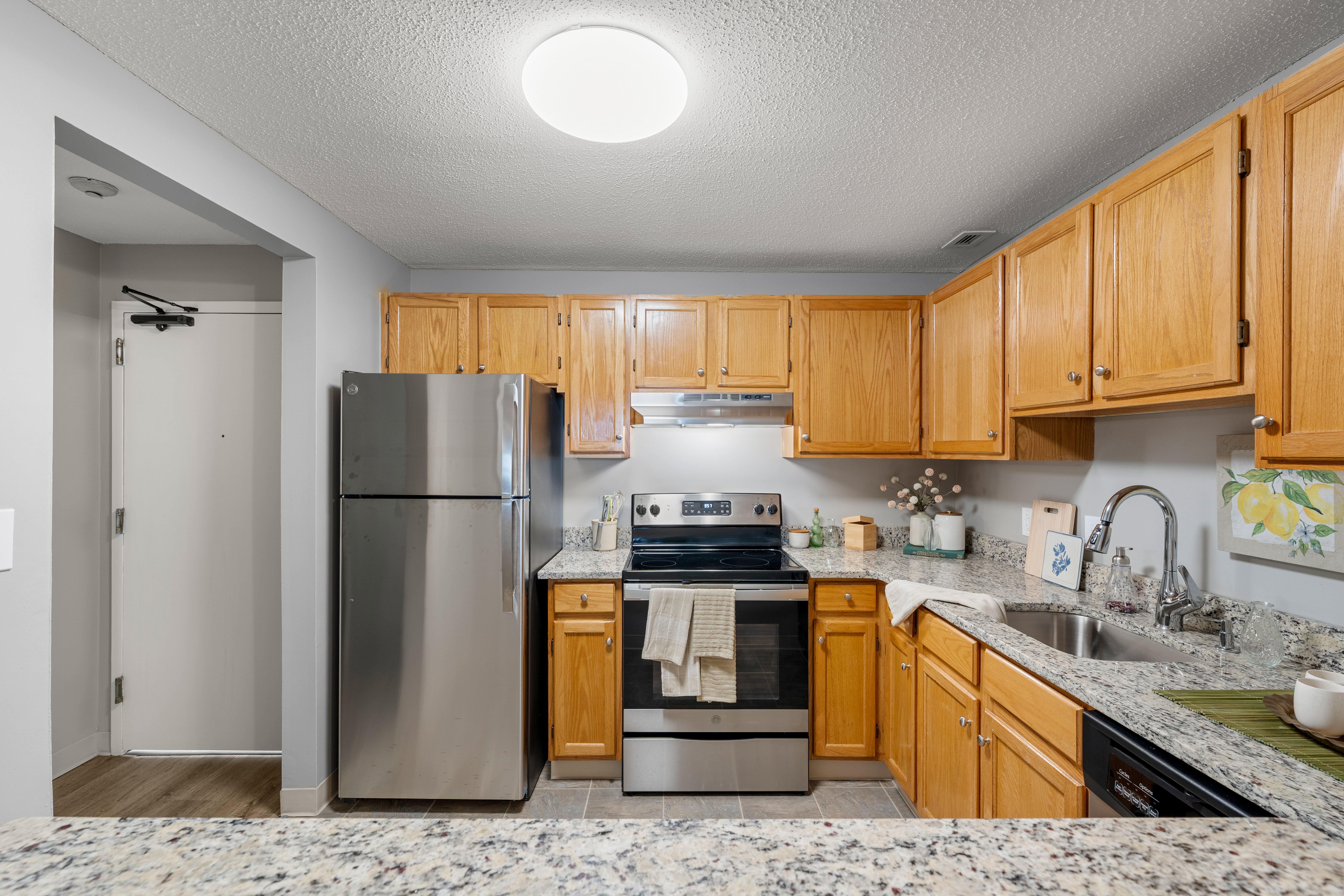 Modern Kitchen Interior A modern kitchen featuring wooden cabinets, stainless steel appliances, and a granite countertop. The kitchen includes a stove, refrigerator, and a sink with a view of the entry door to the left.