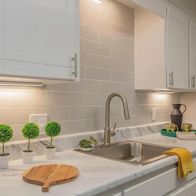 Modern kitchen with white cabinets, a marble countertop, small potted plants, and under-cabinet lighting. Sink and dishwasher visible.