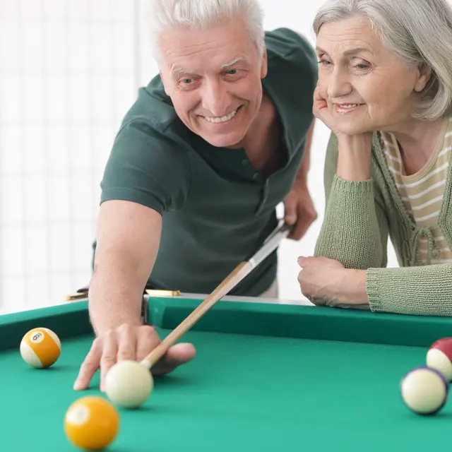 A senior man and woman enjoying a game of pool. The man is smiling and leaning over the table to make a shot, while the woman is watching with an amused expression.