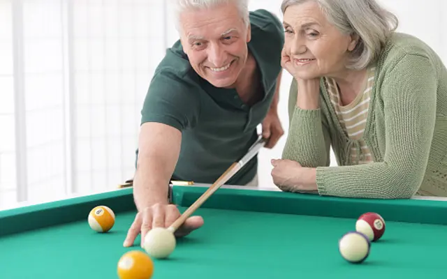 Elderly Couple Playing Pool An elderly man and woman are playing pool. The man is leaning over the green pool table, aiming at a white ball with a cue stick, while the woman watches him fondly, resting her chin on her hand.