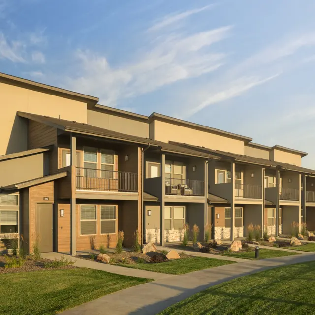 A modern apartment building with multiple units, featuring two stories and balconies, set against a clear blue sky with wispy clouds. The surrounding area includes green grass and landscaped gardens.