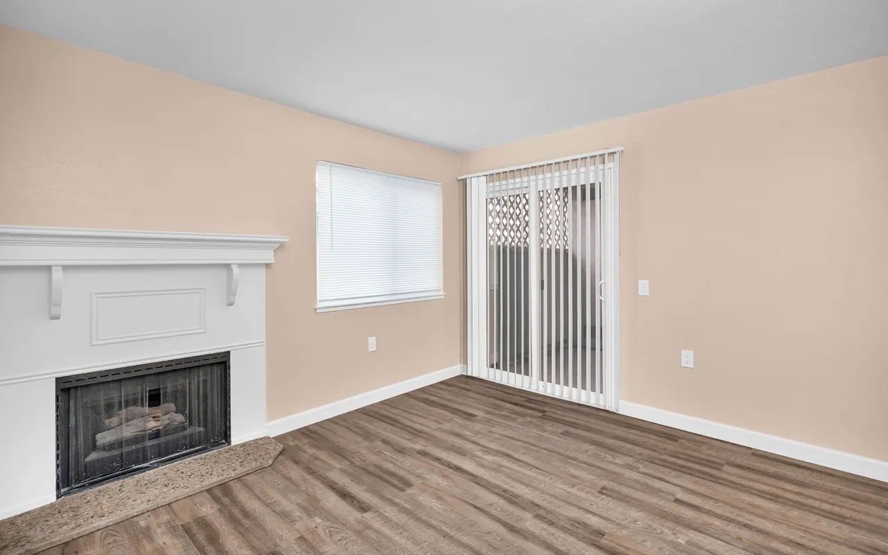 A spacious living room with a fireplace and sliding door leading to a balcony, featuring light-colored walls and wooden flooring.