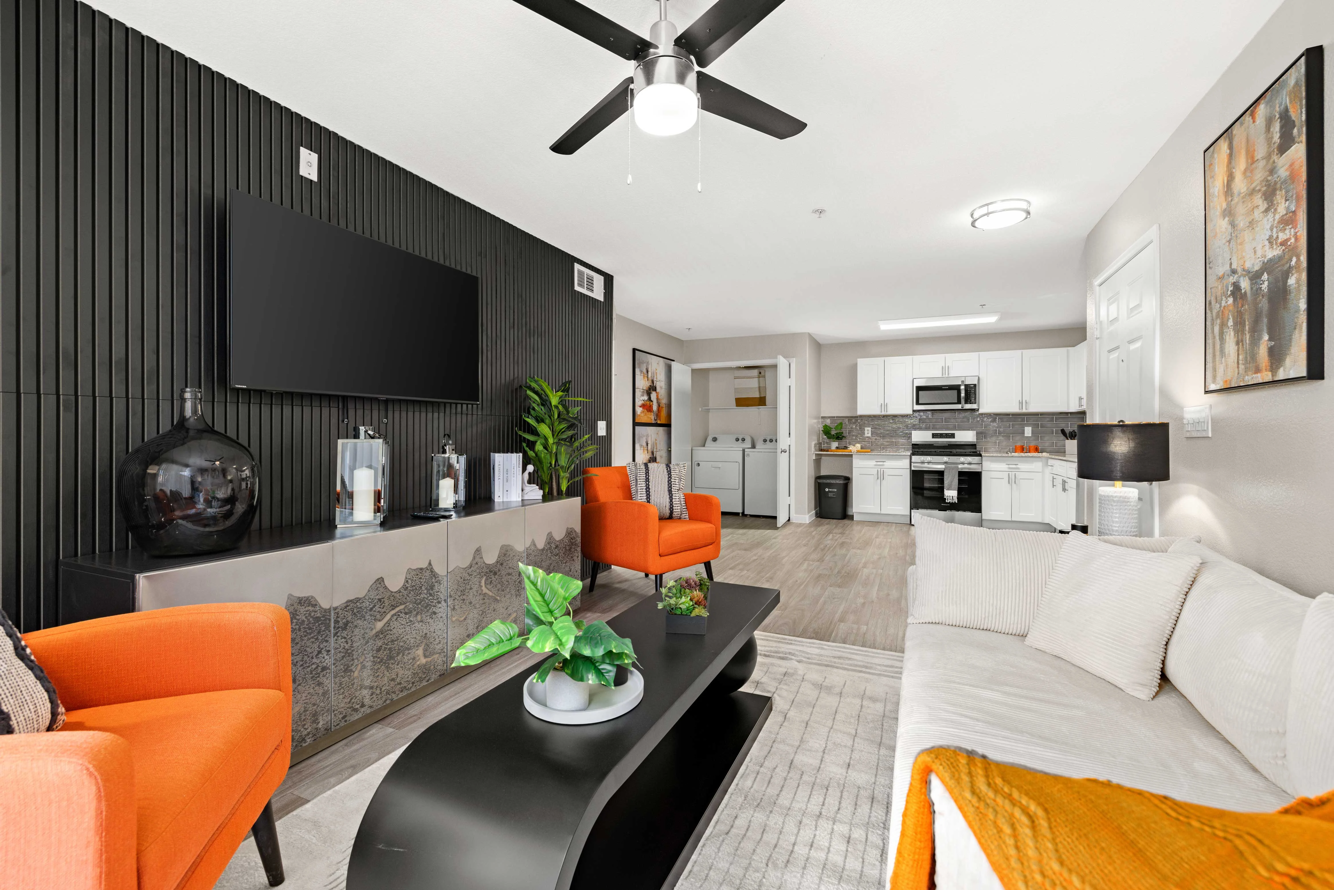 A modern living room featuring a white sectional sofa with an orange throw, an orange accent chair, and a sleek black coffee table. The backdrop includes a flat-screen TV mounted on a textured dark wall, with a small plant near the TV and a bright kitchen visible in the background.