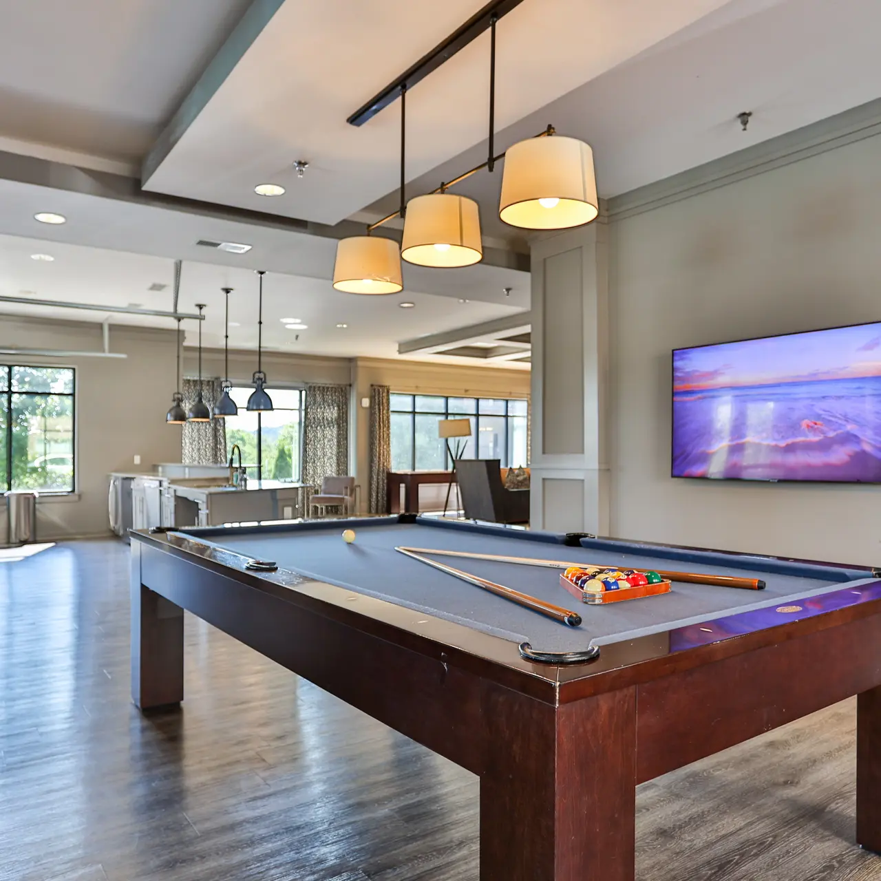 Modern Game Room with Billiards Table A modern game room featuring a billiards table, with two cue sticks and balls on the table. The space is bright and airy, with large windows and pendant lighting. A flat-screen TV is mounted on the wall, displaying a scenic image. There are high chairs lined up against the wall and an open area in the background.