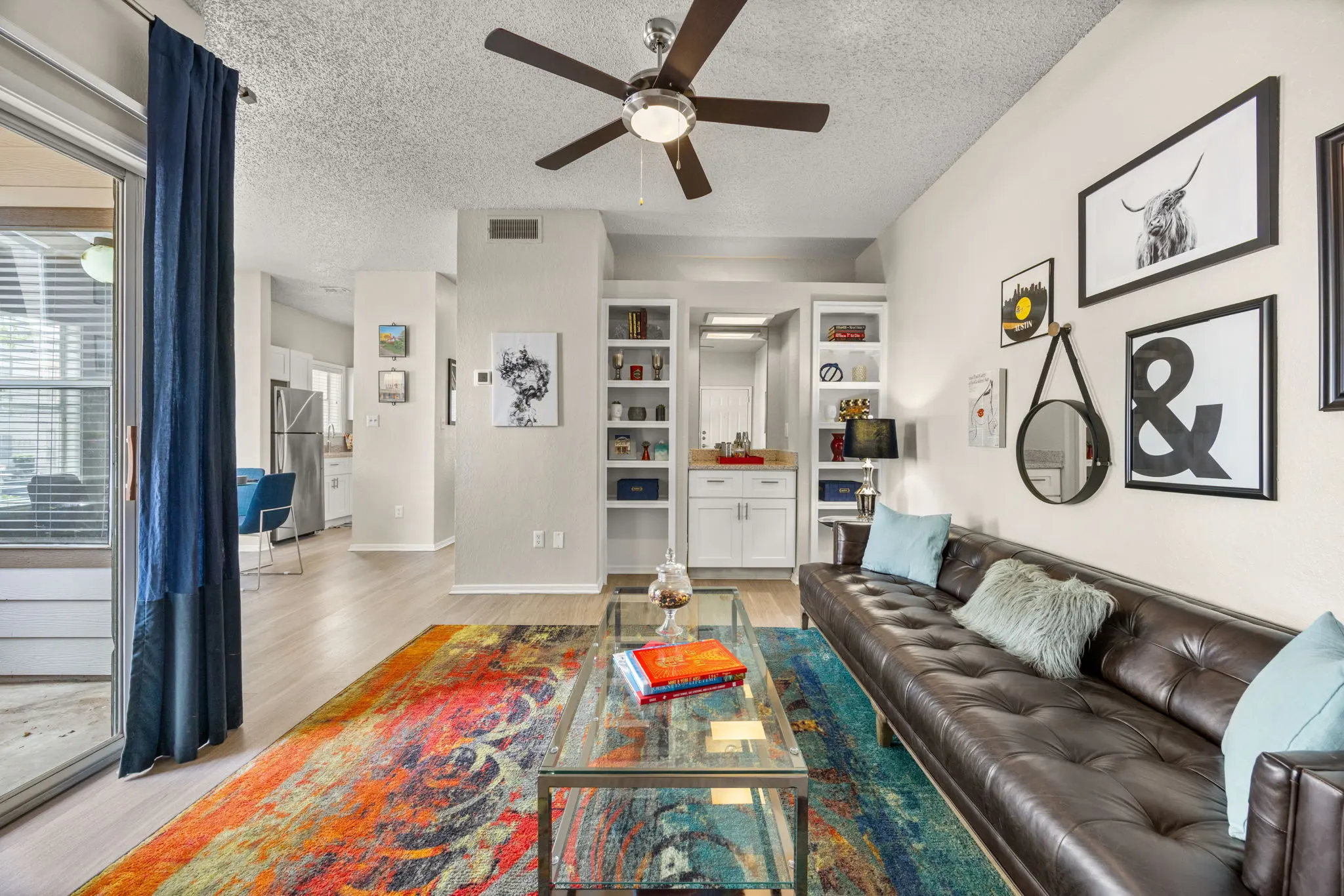 Modern Living Room Design A cozy modern living room featuring a dark brown leather sofa with colorful cushions, a glass coffee table, and a vibrant area rug. The walls are adorned with framed art, and a ceiling fan is visible. In the background, a bright dining area can be seen through an open space with a sliding door. A blue curtain partially frames the window.
