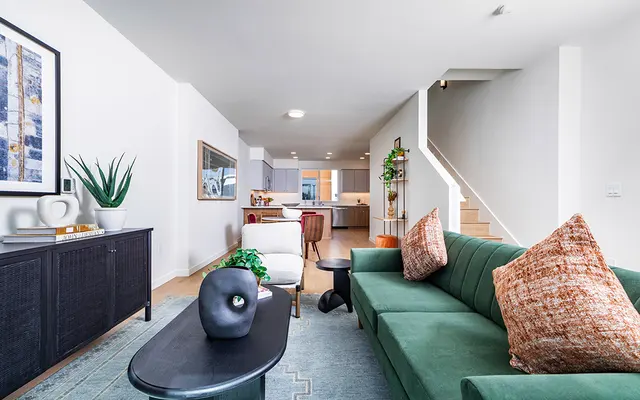 Modern Living Room Design A contemporary living room featuring a green couch with decorative pillows, a black coffee table, and a side chair. The room is well-lit with a minimalist design, including artwork on the walls and plants for decoration. A staircase is visible in the background.