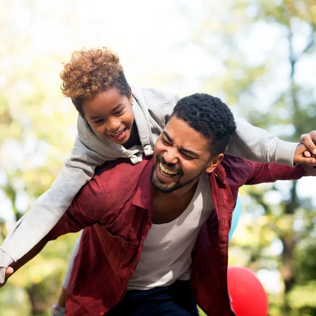 A joyful father giving his son a piggyback ride outdoors, surrounded by greenery and warm sunlight.