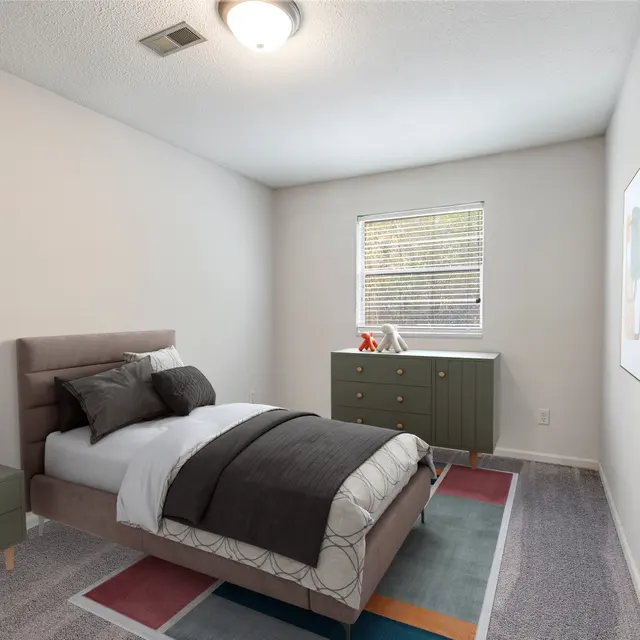 A modern bedroom with a single bed, decorative grey blanket and pillows, small nightstand with a lamp, and a chest of drawers. The walls are white and there are two abstract paintings on the wall. A window provides natural light to the room, and a colorful rug is placed on the carpeted floor.