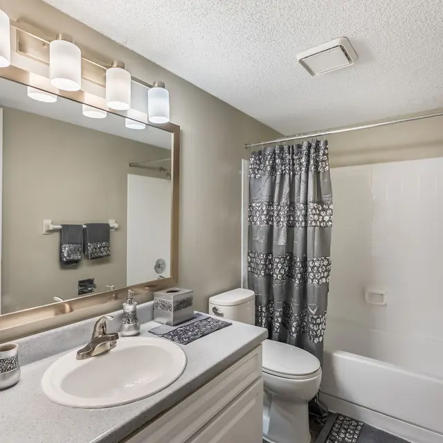 A modern bathroom featuring a white countertop with a sink, a large mirror with light fixtures above it, a gray and white patterned shower curtain, and a clean bathtub.