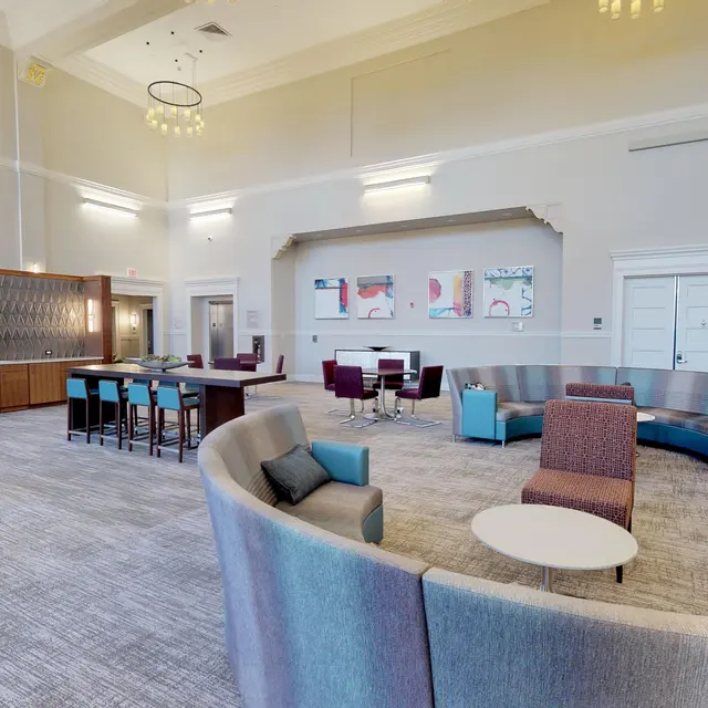 A spacious modern lounge area featuring a variety of seating arrangements, including a curved sofa and chairs. The decor is contemporary with warm lighting and a subtle color palette.