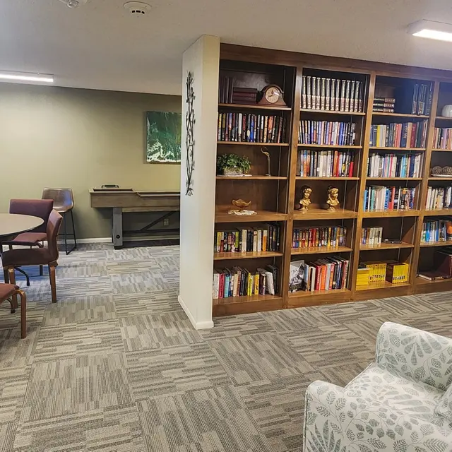 A cozy living space featuring a bookshelf filled with books, a table with chairs, a comfortable couch, and a piano in the background.