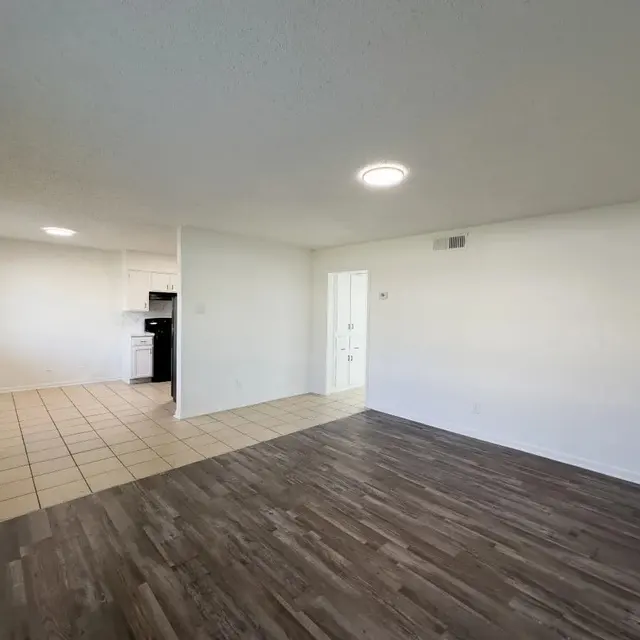 Spacious Living Area A spacious living area featuring a combination of tiled and wooden flooring. On the left, there is a doorway leading to a kitchen with black appliances. The walls are painted white, and there is overhead lighting.