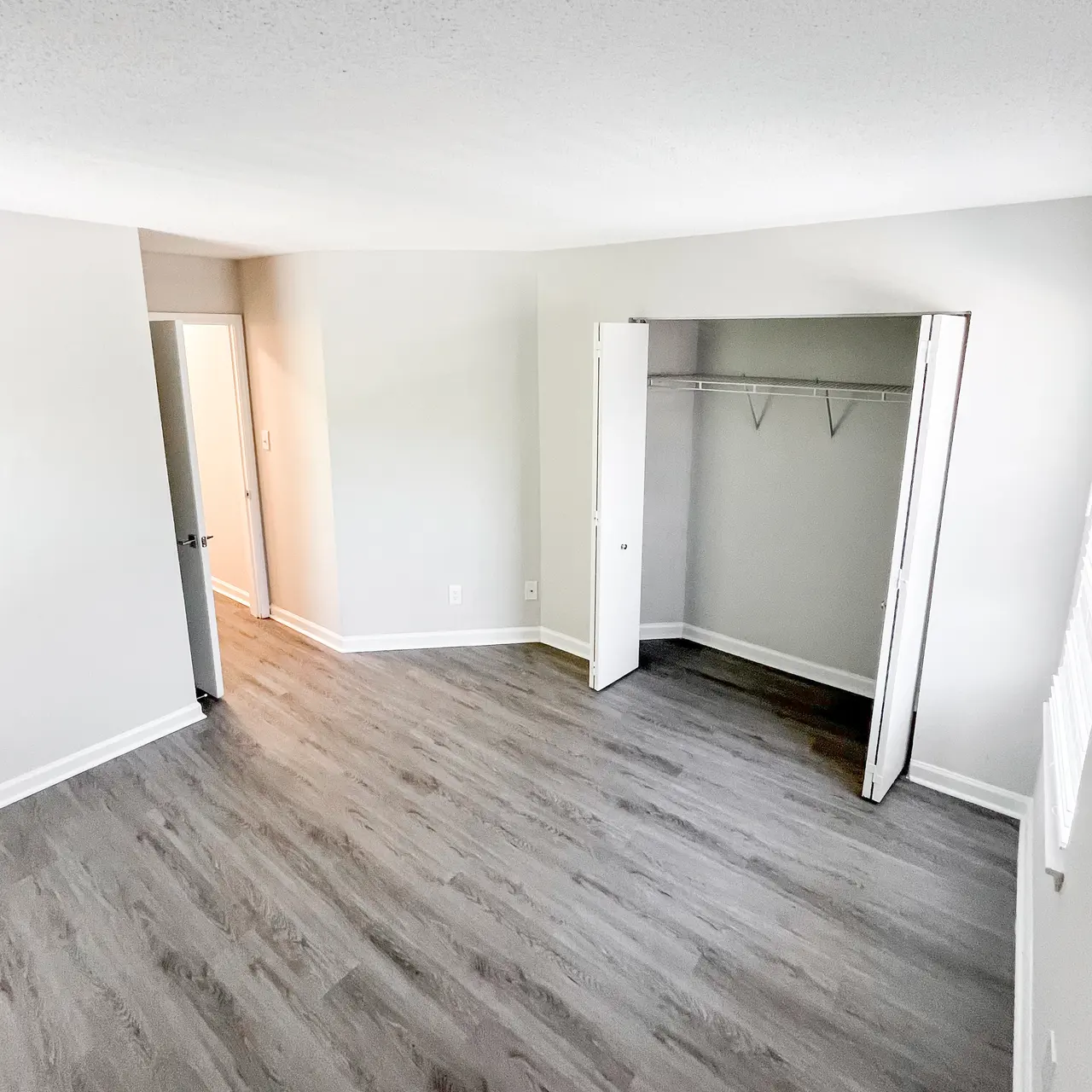 Spacious Empty Room A spacious empty room with light gray walls and light wood flooring. A closet with open sliding doors is visible on the right, and a doorway leading to another room is seen on the left.