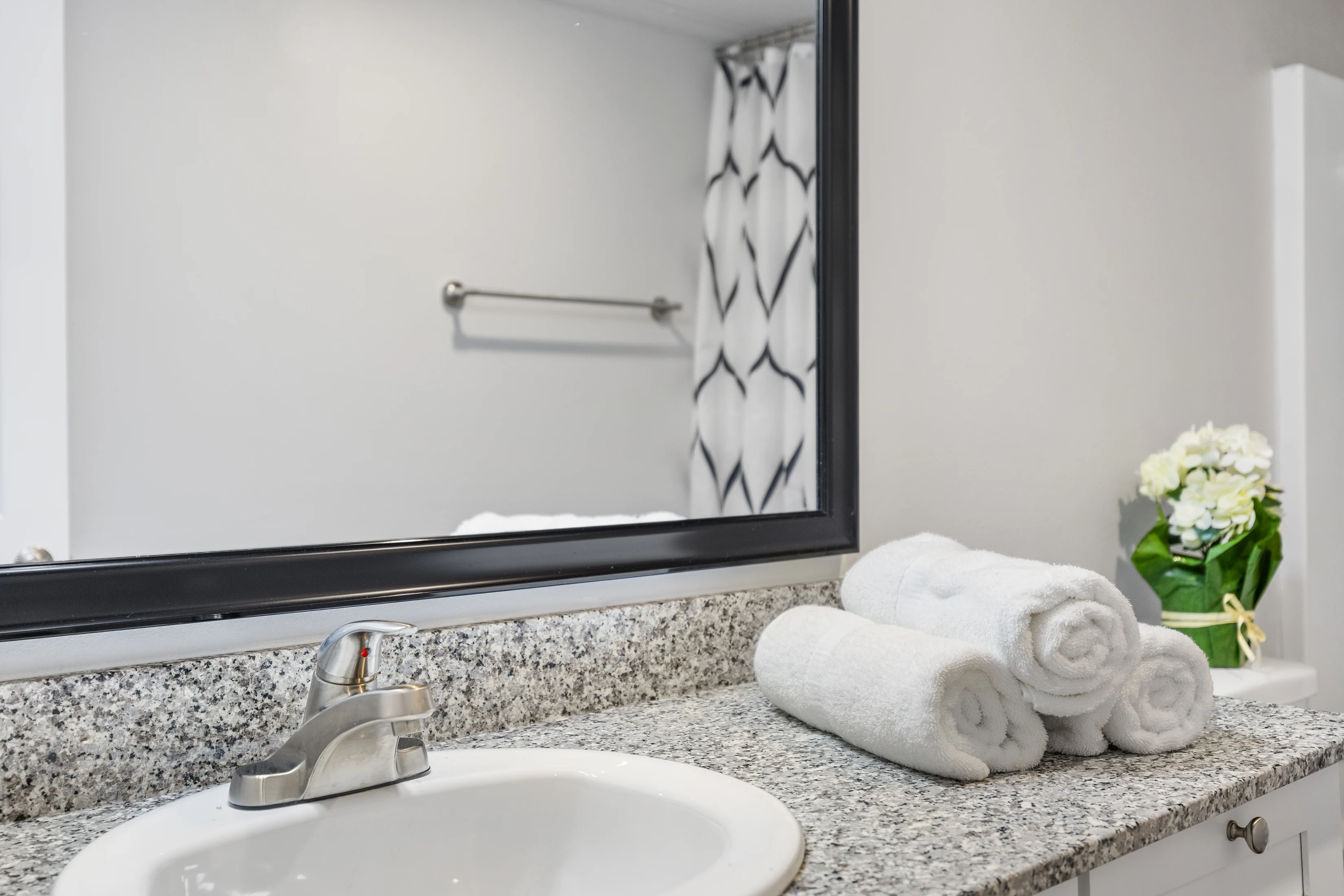 Modern Bathroom Sink View A modern bathroom sink with a faucet on a granite countertop, neatly rolled white towels next to it. A mirror reflects the minimalistic decor, including a plant arrangement in the background.