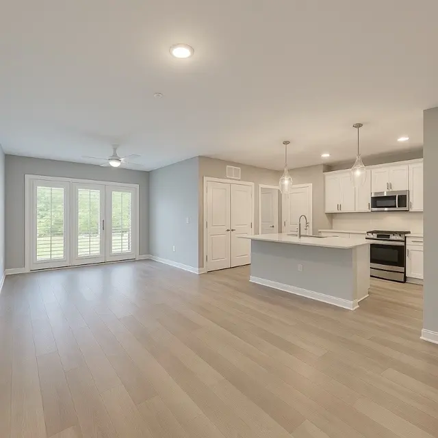 Contemporary Living Space A spacious modern living area with wooden flooring and a light gray color scheme. The room includes double doors leading to an outside deck, a ceiling fan, and a kitchen with white cabinets and stainless steel appliances.
