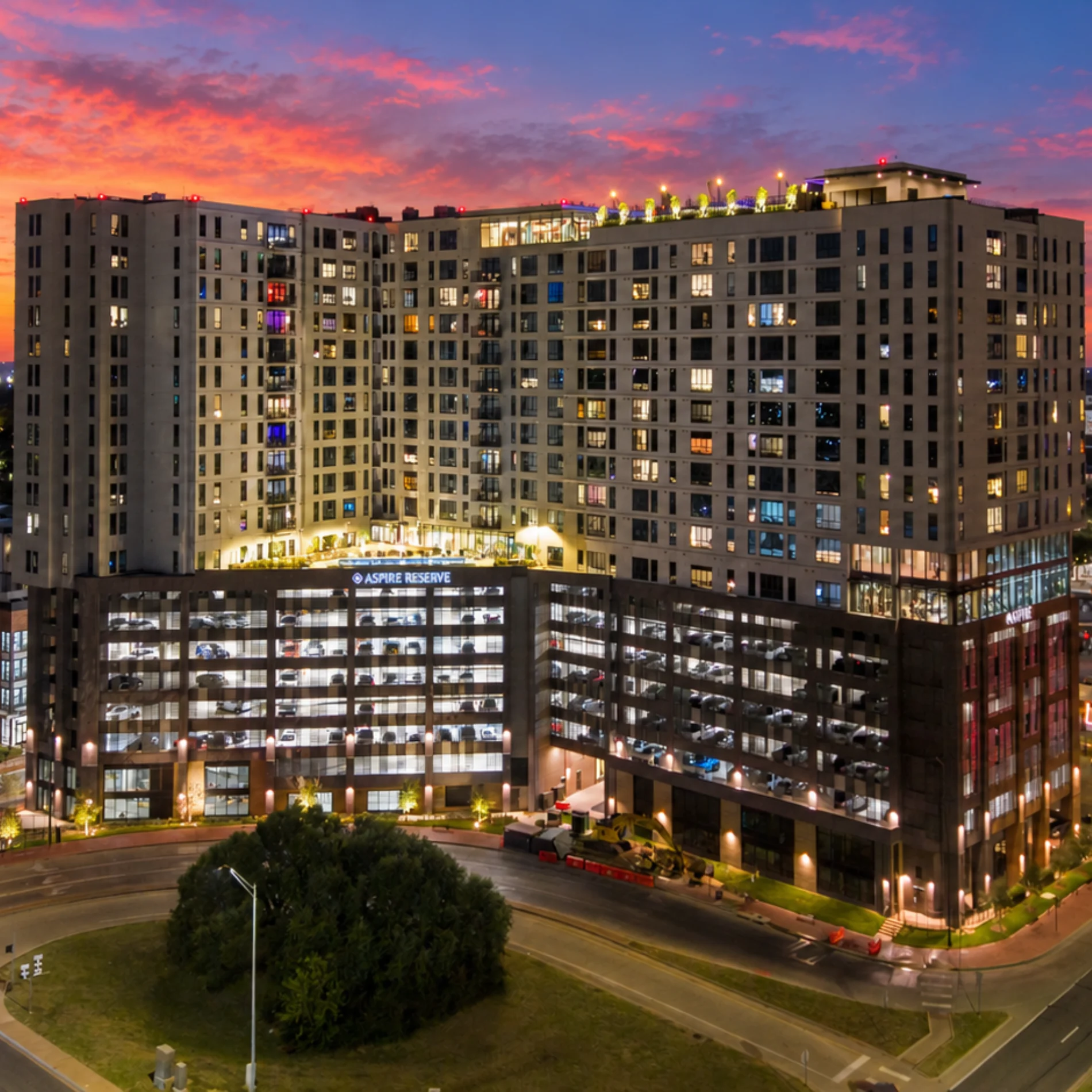 A modern apartment complex illuminated at sunset, featuring multiple floors with balconies and a parking garage.