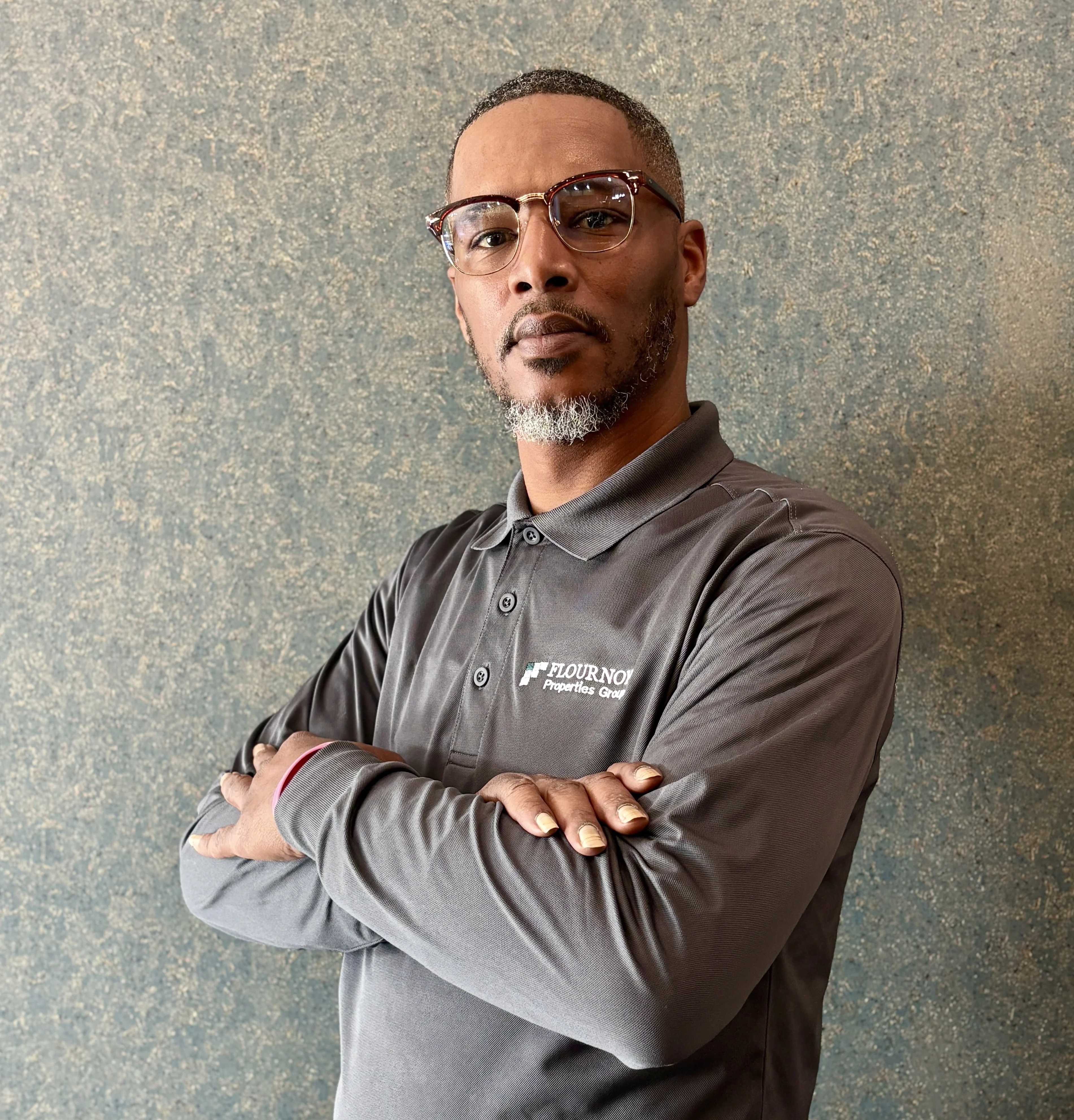 Professional Man with Glasses A confident man in a gray polo shirt with glasses, standing with crossed arms against a textured blue background.