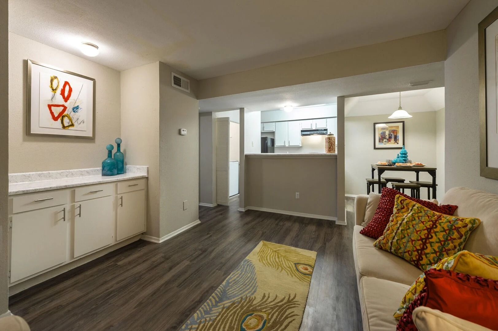 Interior view of a modern apartment living room with a sofa, decorative pillows, and a rug, leading to a kitchen area in the background.