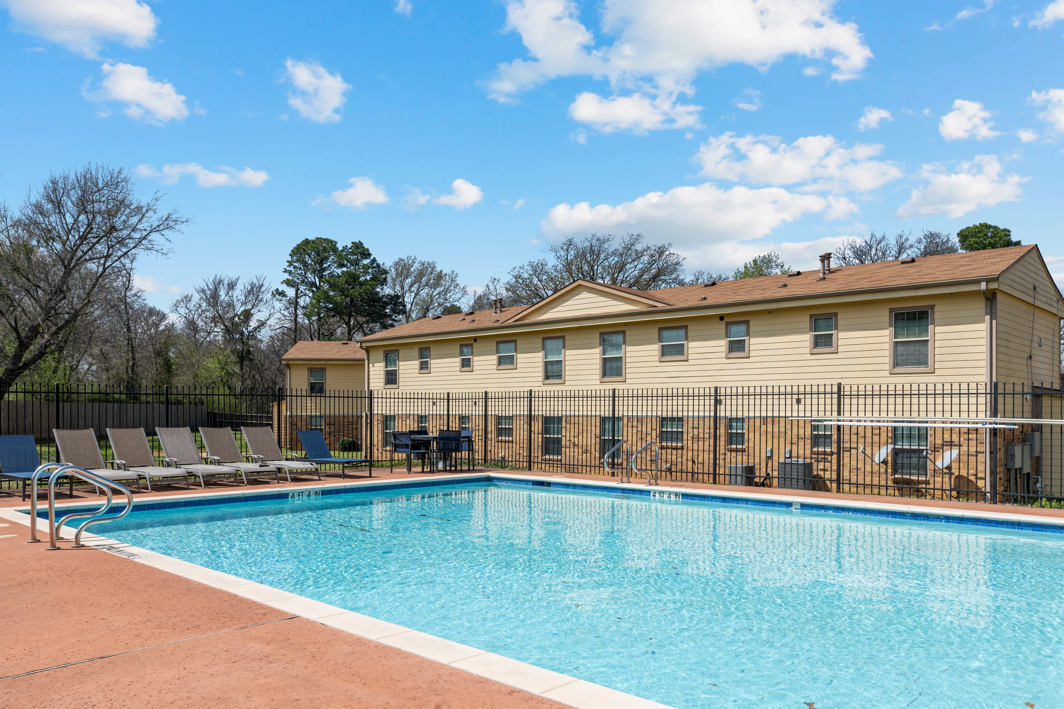 Apartment Complex Pool Area A clear swimming pool surrounded by lounge chairs with an apartment building in the background, under a blue sky with fluffy clouds.