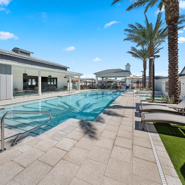 A sparkling pool surrounded by palm trees, lounge chairs, and modern buildings under a clear blue sky. 