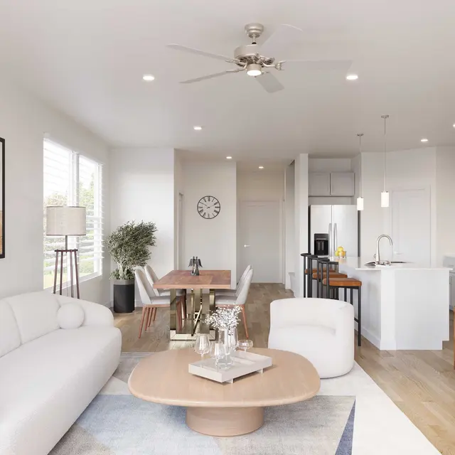 A stylish modern living room featuring a large white sofa, a round coffee table, and a wooden entertainment unit with a flat-screen TV. The space has bright lighting, light wooden flooring, and a dining area with a wooden table and chairs in the background.