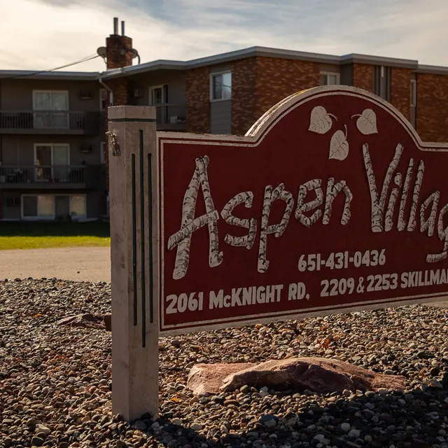 Sign for Aspen Village with contact information and address details, in front of an apartment building.