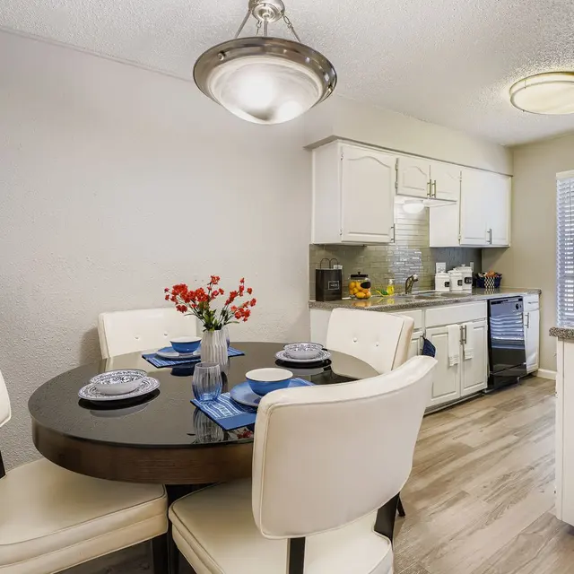 Modern Kitchen and Dining Space A modern kitchen area featuring a round dining table with four white chairs, plates, and a vase of red flowers. The kitchen is equipped with white cabinets and stainless steel appliances, with sunlight streaming through a window.