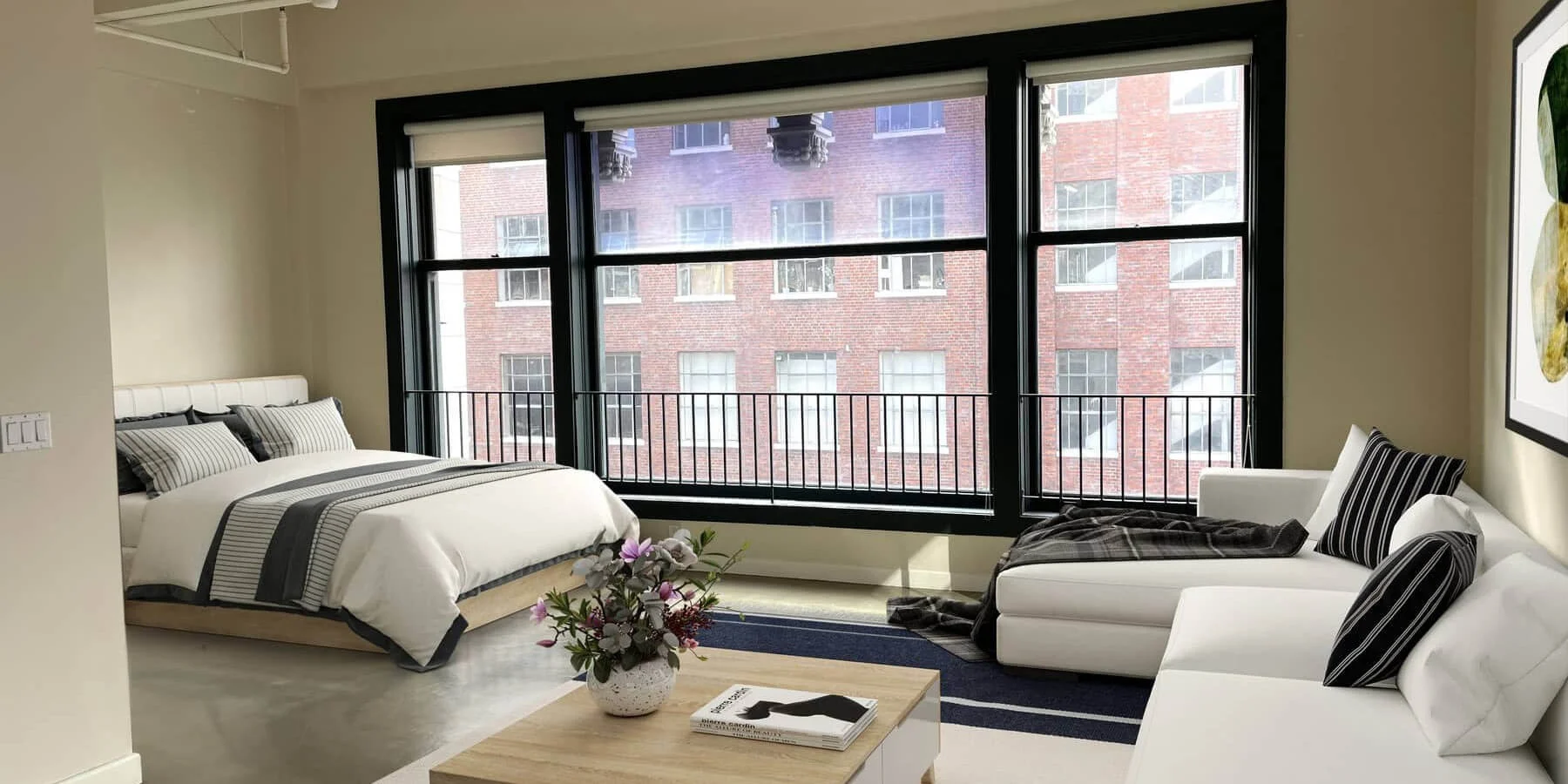 A modern studio apartment with large windows, featuring a bed and a sofa, with minimalistic decor and a small table with flowers.