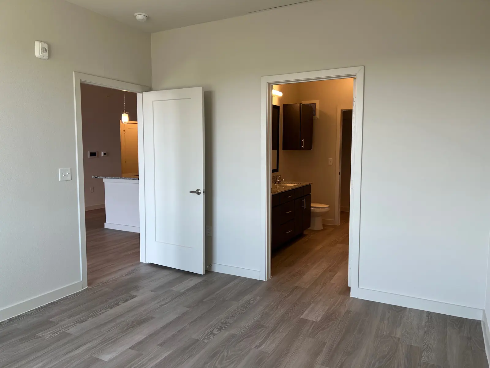 A view from a spacious room with light-colored walls, showcasing a doorway that leads to a bathroom area and another door leading to a kitchen or living space. The floor is made of light wood, creating a modern aesthetic.