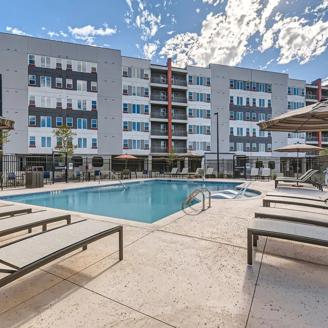 A modern apartment complex with a swimming pool surrounded by lounge chairs and umbrellas under a partly cloudy sky.