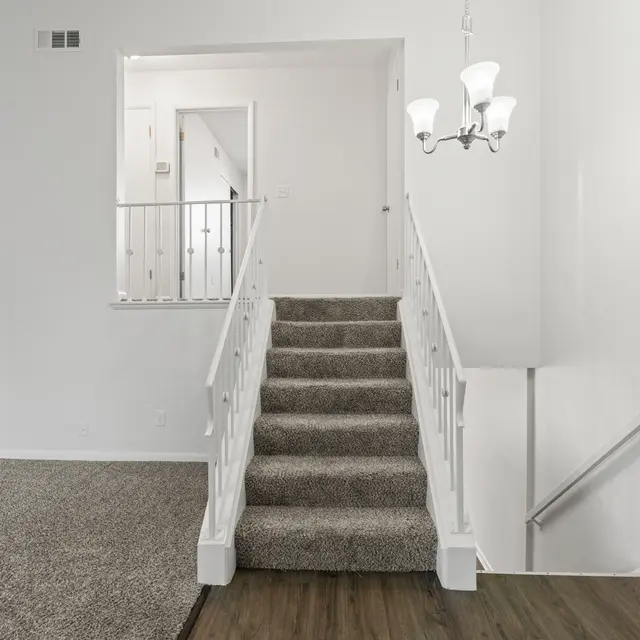 A view of the interior of a home showcasing a staircase leading to an upper level. The staircase has beige carpet and white railings. To the left, there is a view into a room through an open door, revealing light-colored walls and a wood floor. The room on the main level has a carpeted area and there is a chandelier hanging from the ceiling.