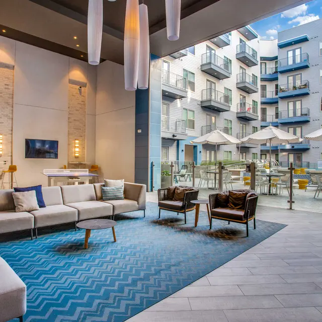 A contemporary lounge area with a large sectional sofa, two chairs, and decorative pillows, featuring a blue zigzag patterned rug. Large glass doors open to an outdoor seating area with umbrellas and an apartment building in the background.