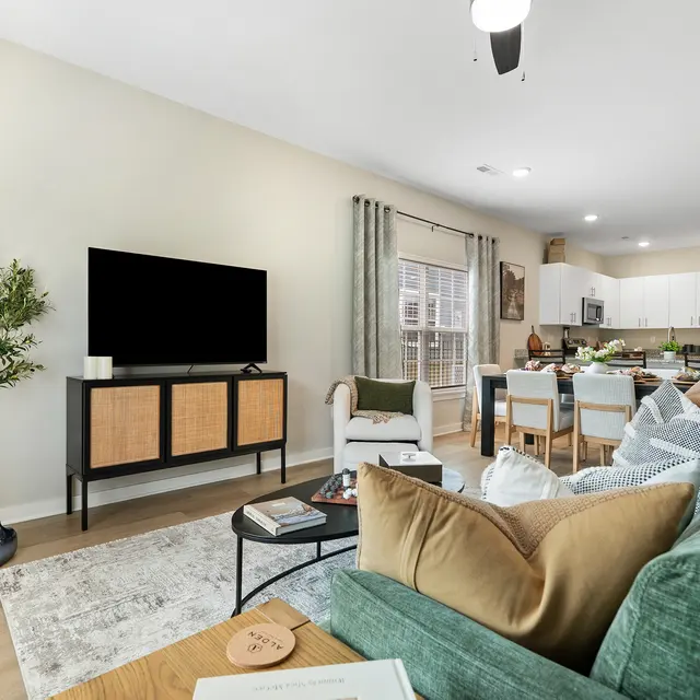 A stylish modern living room featuring a green sofa, wooden coffee table, and a large TV on a stand. There is a dining area in the background with a kitchen, plants, and natural light coming through the windows.