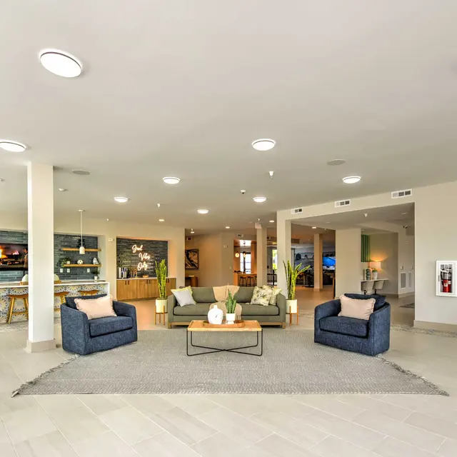 Elegant Lobby Interior A spacious and modern lobby area with two blue armchairs, a coffee table, and a decorative centerpiece on a light gray rug. Background features a counter and shelves with decorative items and plants, along with large light fixtures overhead.