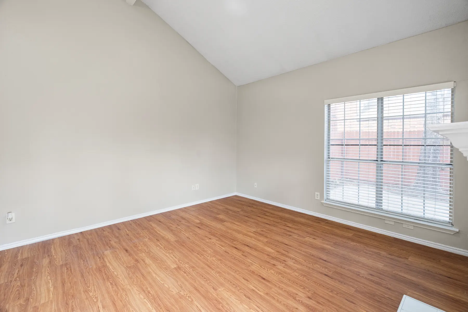 An empty living room with wooden flooring and a vaulted ceiling. A large window with white blinds lets in natural light.