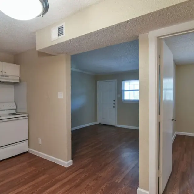 Interior view of a small apartment featuring a kitchen area with a stove, refrigerator, and brown wooden flooring. A doorway leads to a living room area with a front door and windows.