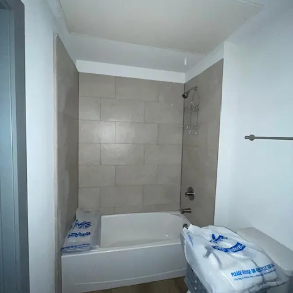 A minimalist bathroom featuring a bathtub, shower fixture, and a toilet. The walls are tiled with light-colored tiles, and there are items stacked near the bathtub.